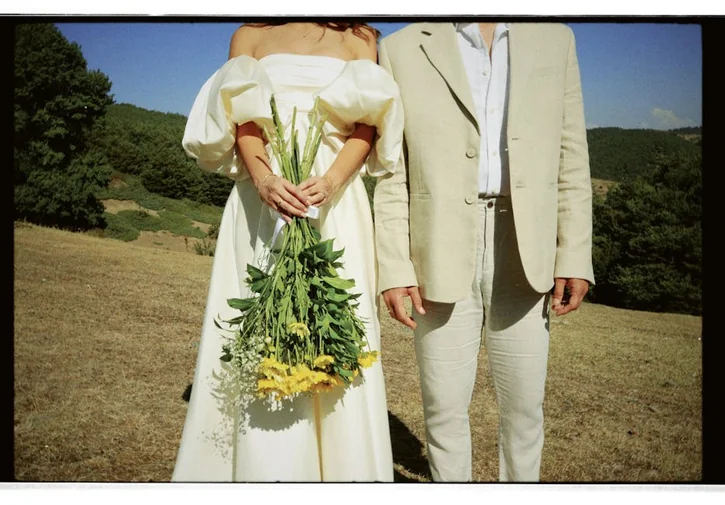 A couple dressed in wedding attire stands in an outdoor setting, holding a bouquet.