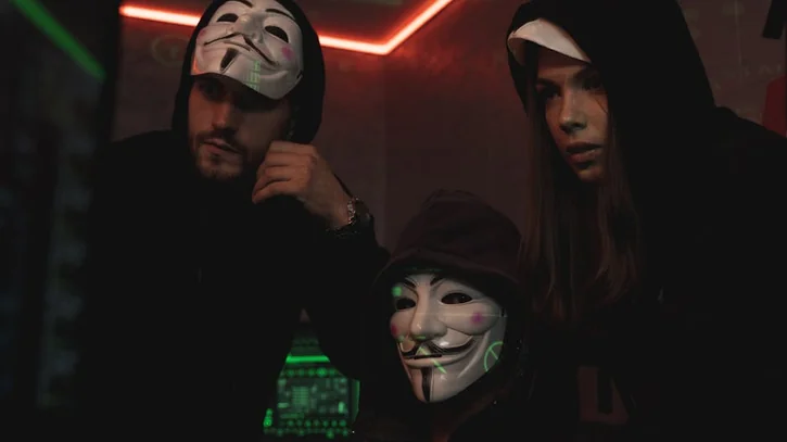 A group of people wearing Guy Fawkes masks in a dark tech-themed room, suggesting anonymity and hacking activities.