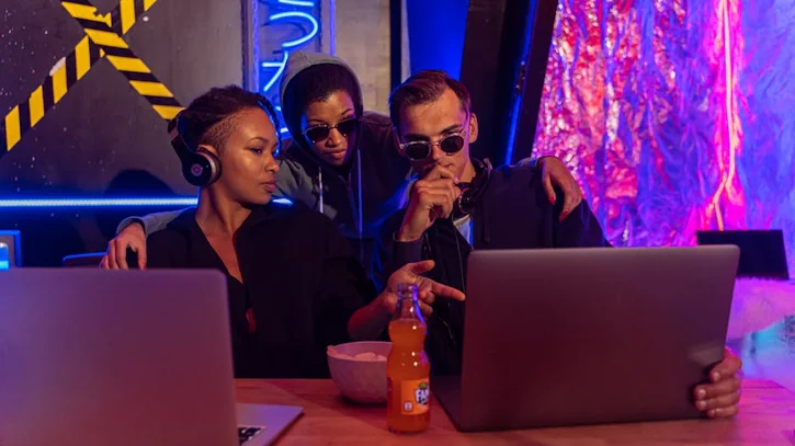 Three people collaborate on laptops in a cyber-themed workspace, discussing strategies.