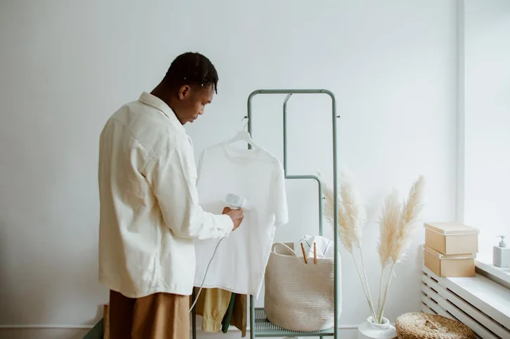A man steams a white t-shirt on a clothing rack indoors, showcasing a minimalist style.