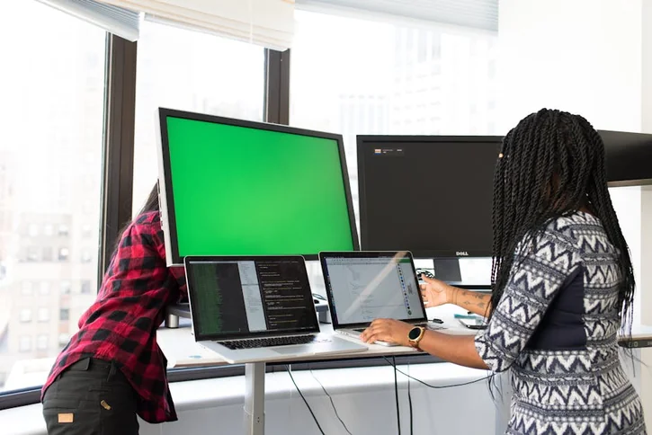 Two women working on laptops and monitors in a bright office setting, focused on technology and teamwork.