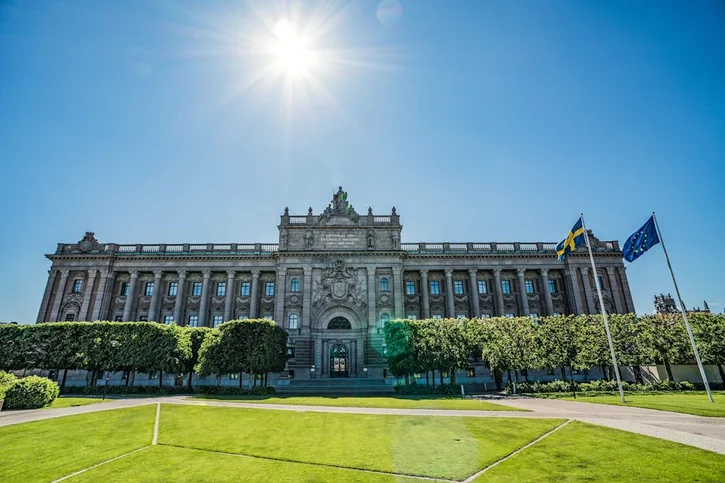 Front view of the Riksdag building with Swedish and EU flags on a sunny day.