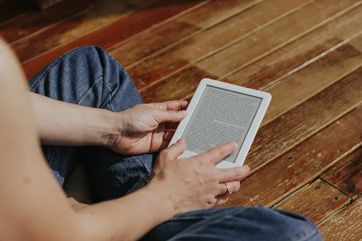 Close-up of hands holding an e-reader indoors on a wooden floor, emphasizing digital reading.