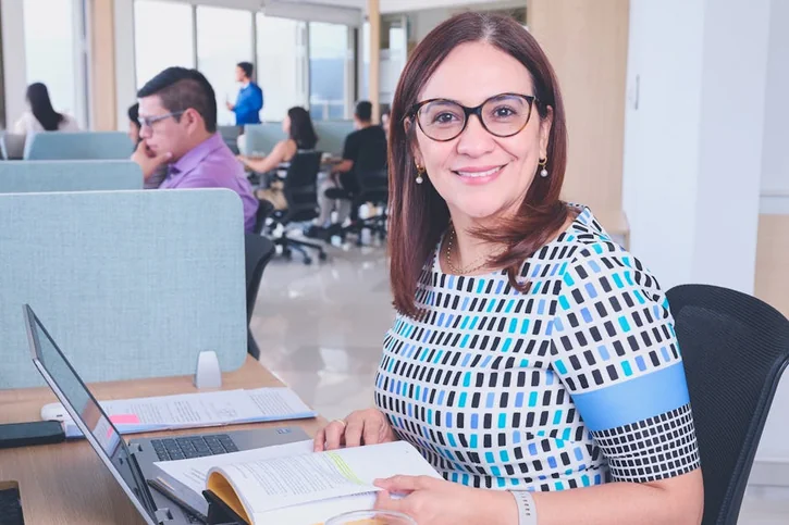 Confident woman in a modern office, surrounded by colleagues, working on a laptop.