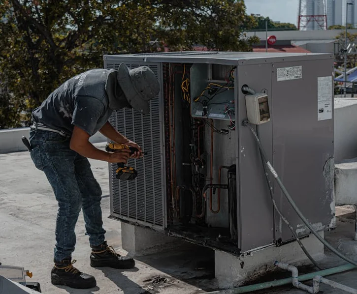 A worker in a bucket hat repairs an outdoor air conditioning unit on a rooftop.