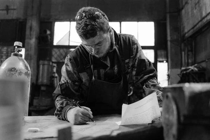 Blacksmith in a workshop drawing plans with concentration and precision, surrounded by tools and machinery.