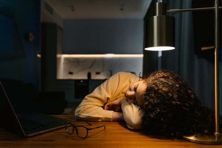 Exhausted woman sleeps at her desk under a lamp with a laptop beside her.