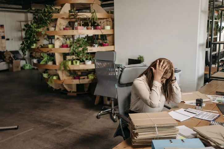 A stressed woman sits overwhelmed at her desk, surrounded by paperwork in a modern office setting.