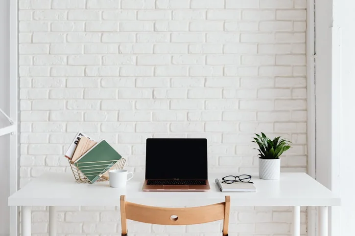 Clean and organized white workspace with laptop, plant, and books against a brick wall.