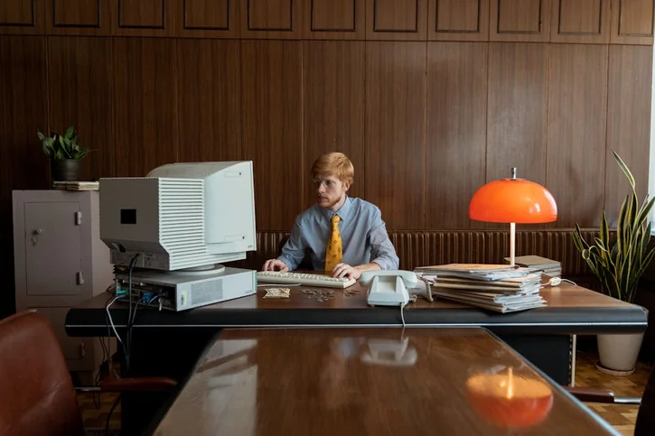 Vintage office environment with a man working on a classic CRT monitor and desktop computer setup.