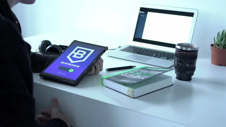 ブロックチェーンを表示するタブレット、ノートPC、本、カメラレンズが置かれたデスク