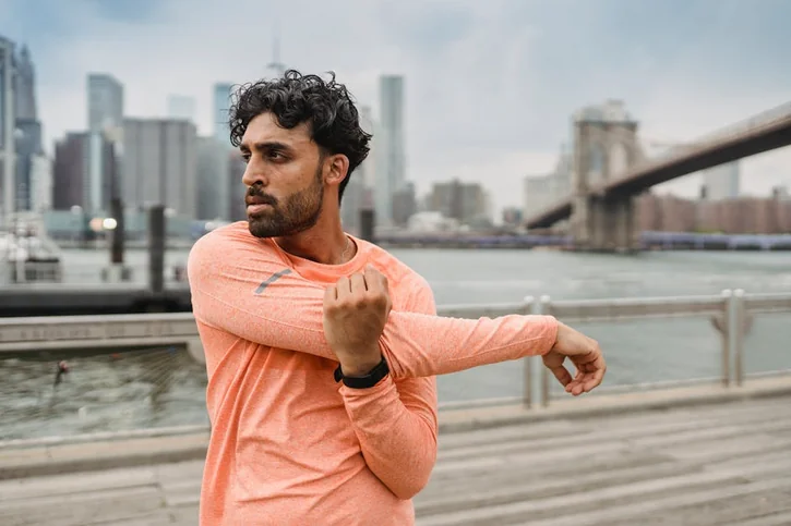 Man in orange sweater stretching by the river with city skyline and bridge in view.