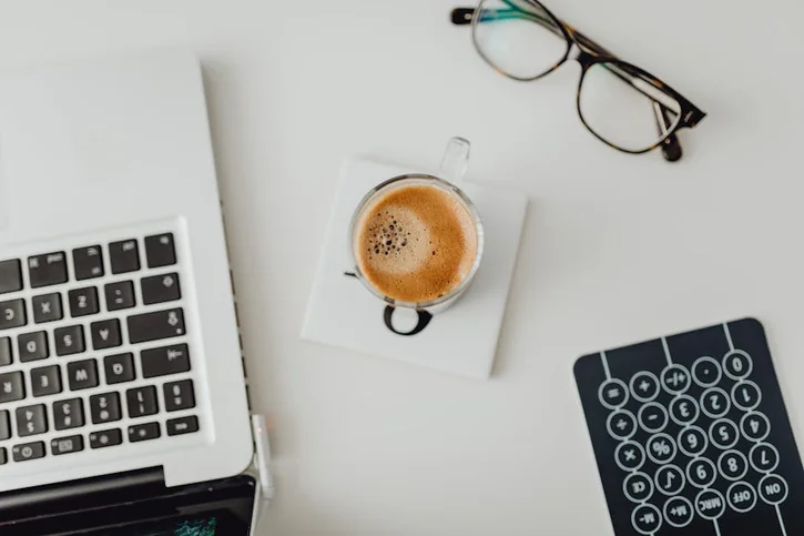 Top view of a modern workspace featuring a laptop, eyeglasses, coffee cup, and calculator on a desk.