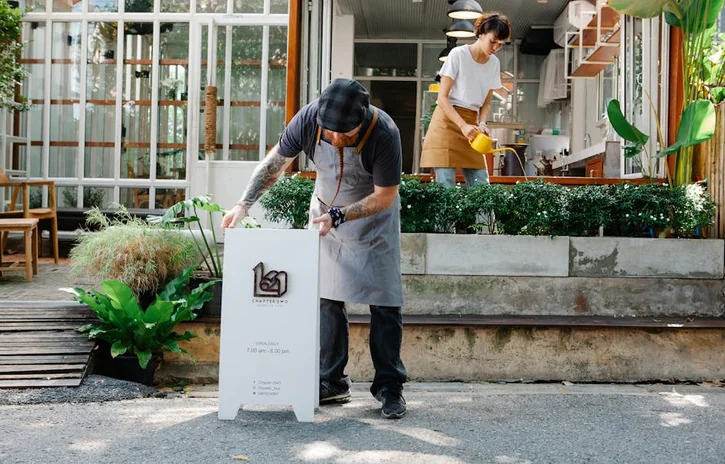 Couple of coworkers in aprons and casual clothes watering plants and putting signboard near cafeteria in daytime