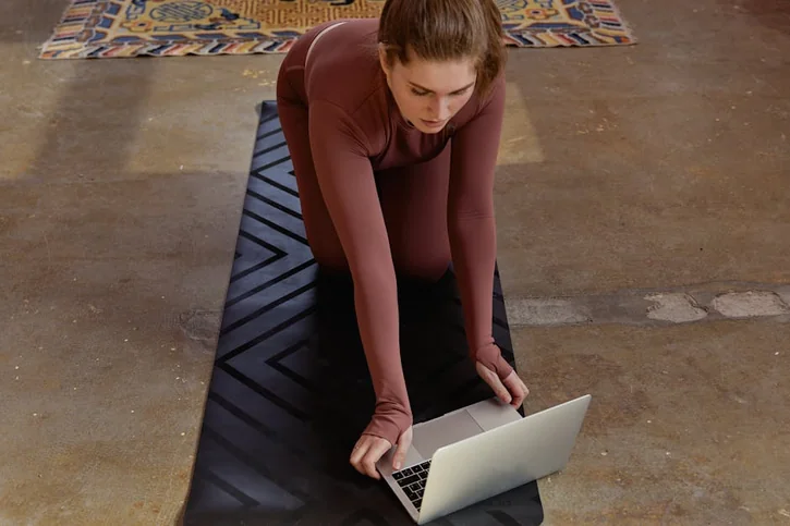Woman in yoga attire kneeling on mat using laptop indoors, blending fitness with technology.
