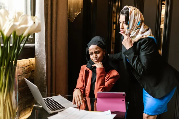 Two women in business attire discuss work at a laptop in a modern office.