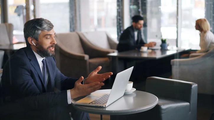 A businessman in a café video conferencing on a laptop using wireless headphones.
