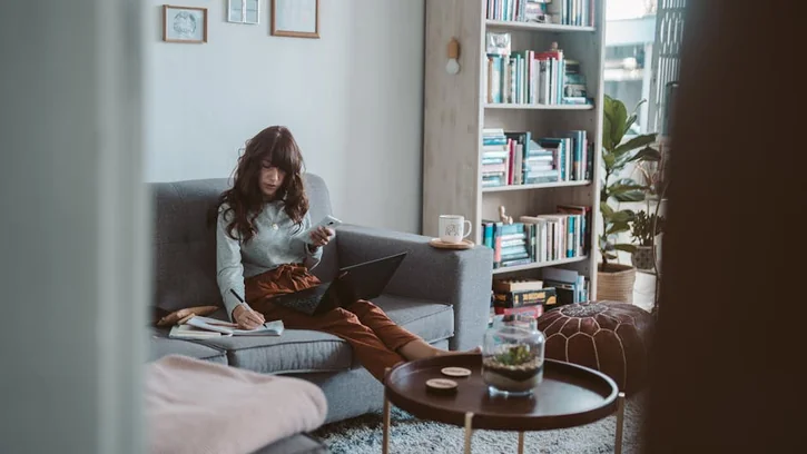 Woman seated on a sofa working remotely with a laptop and phone in a cozy home setting.