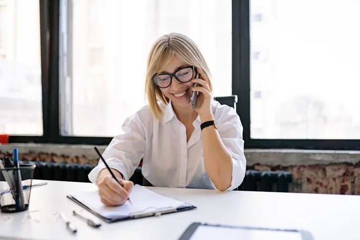 Smiling woman talking on the phone while writing notes at a desk in a modern office.