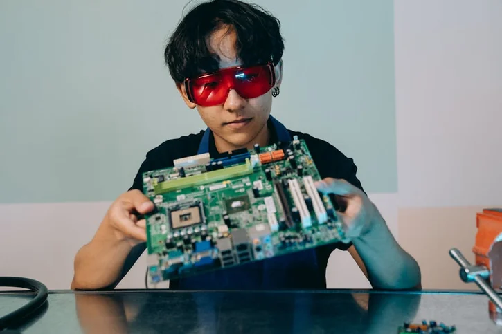 A focused young engineer holding a motherboard, wearing eye protection indoors.